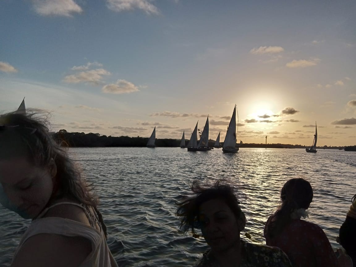 Uitzicht vanaf een dhow bij zonsondergang: een vloot zeilboten op het water bij Lamu, met op de voorgrond donkere silhouetten van medeopvarenden.