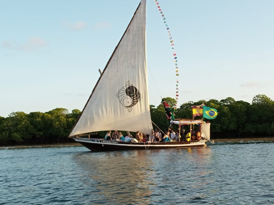 Een dhow onder vol zeil op het water bij Lamu, met een lange vlaggenlijn en mangroven op de achtergrond. Op het zeil een rond teken van twee vissen, een zwarte en een witte, die in elkaar grijpen.